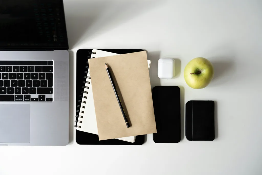 A laptop, notebooks, phone and green apple lined up on a white desk showing someone preparing to work on a cloud engineer resume.