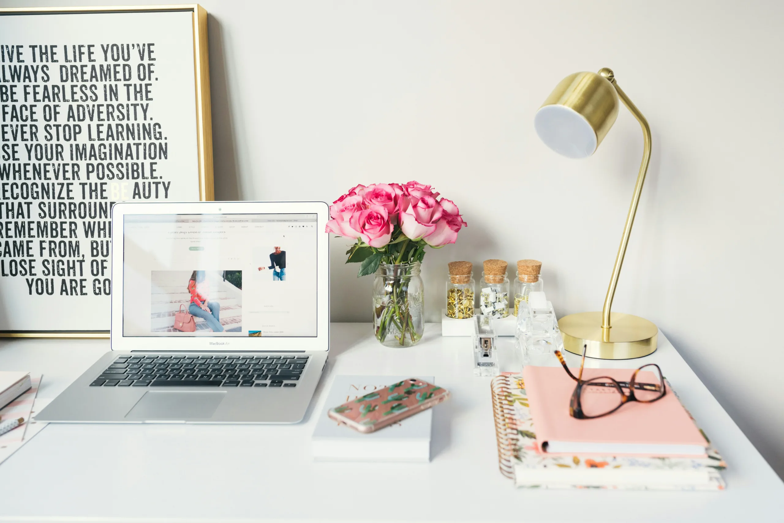 A white desk with a laptop, pink flowers and a gold lamp on it showing the workspace of a tech assistant.