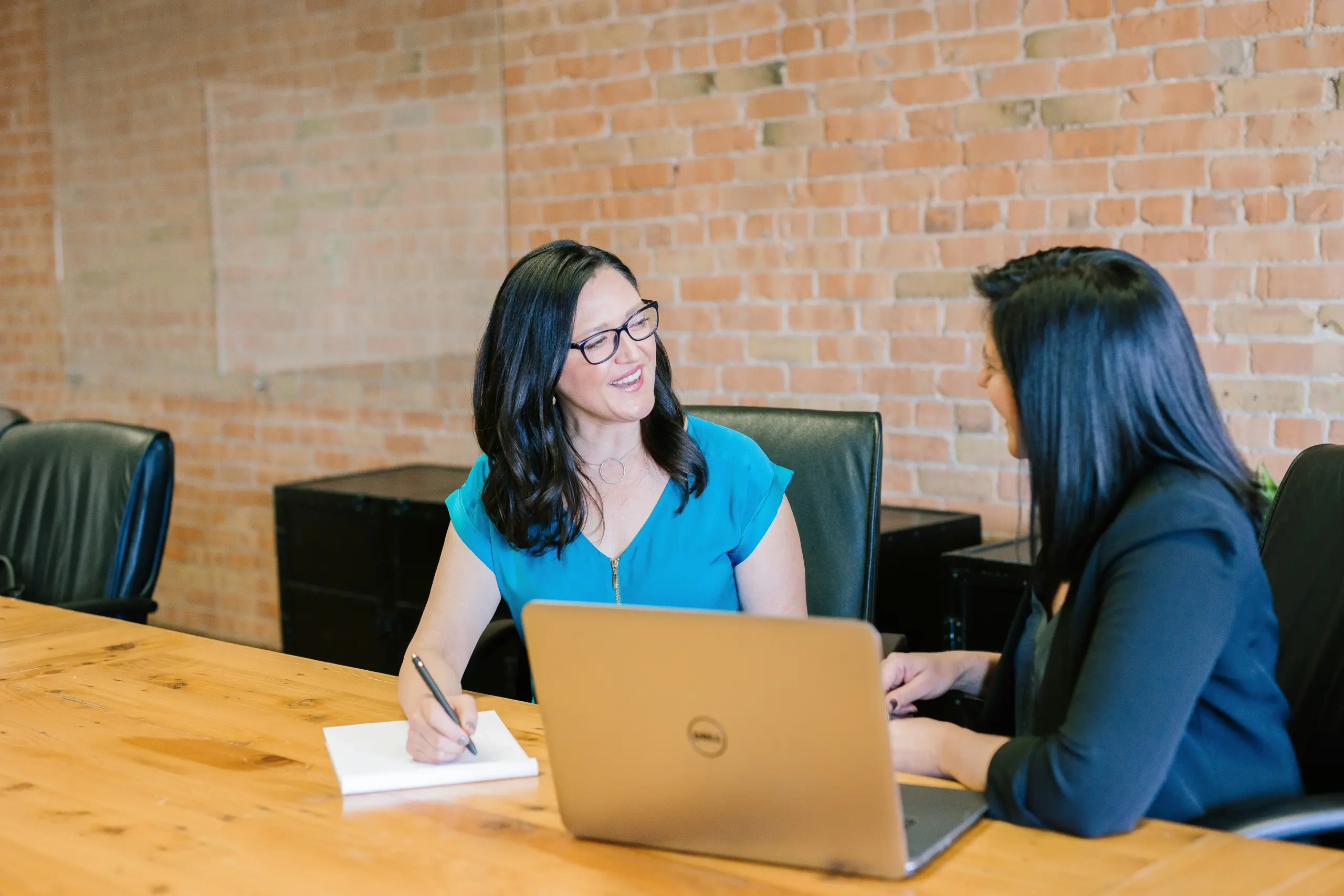 Two women talking at a table with an open laptop in front of them.