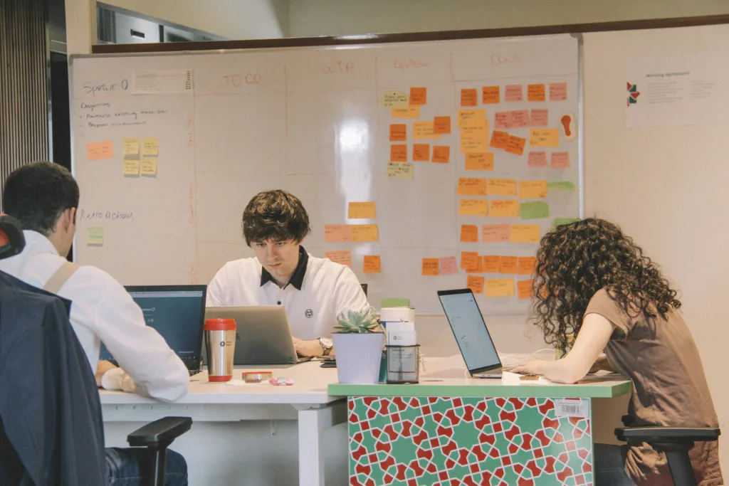 A group of people working at a table in front of an agile board showing which basic agile quality practice reduces bottlenecks and ensures consistency.