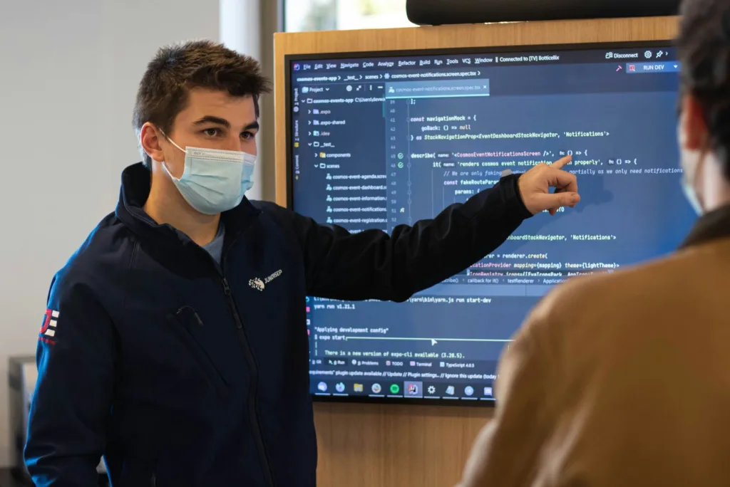 A man with a blue face mask pointing to a board of code at a C# boot camp.